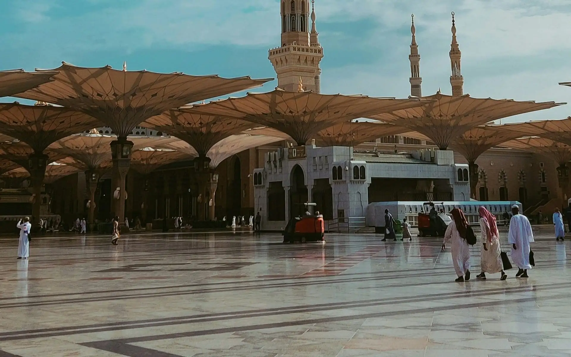 The courtyard of Masjid an-Nabawi in Madinah with its shade canopies open and pilgrims walking at sunrise