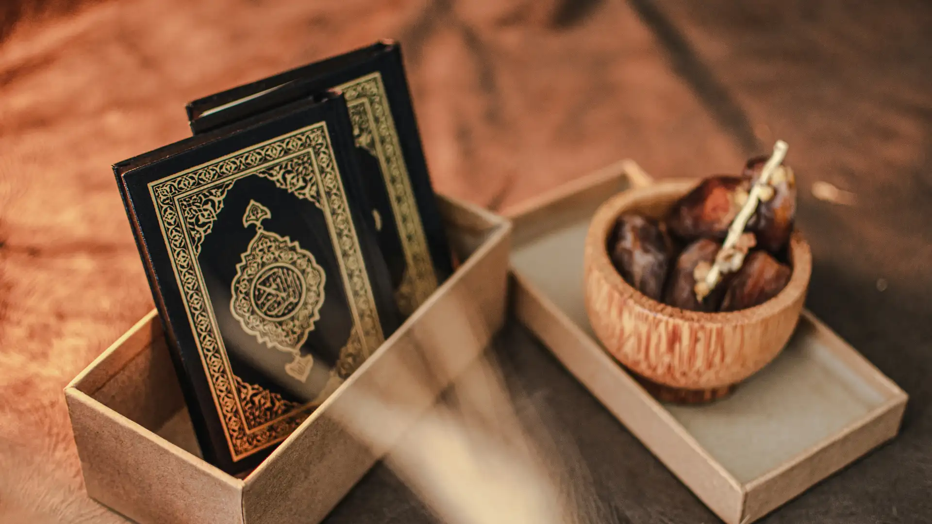 A gold embossed Quran resting in a gift box beside a wooden bowl of dates and stalks of wheat, in warm evening light